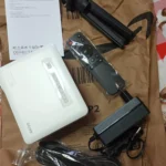 Contenuto della confezione del proiettore 4K economico Akiyo O1: mini proiettore, telecomando, cavi, alimentatore, mini treppiede e manuale, disposti su un imballaggio in carta.