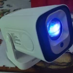 Proiettore 4K economico Magcubic HY310X bianco acceso, con lente illuminata di blu, appoggiato su una superficie domestica.