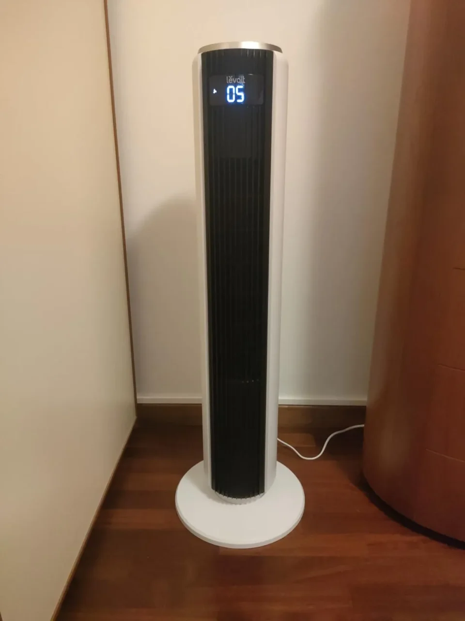 Ventilatore a torre Levoit acceso con display LED frontale su pavimento in legno chiaro, posizionato di fianco a un mobile per mostrare il suo design compatto salvaspazio.